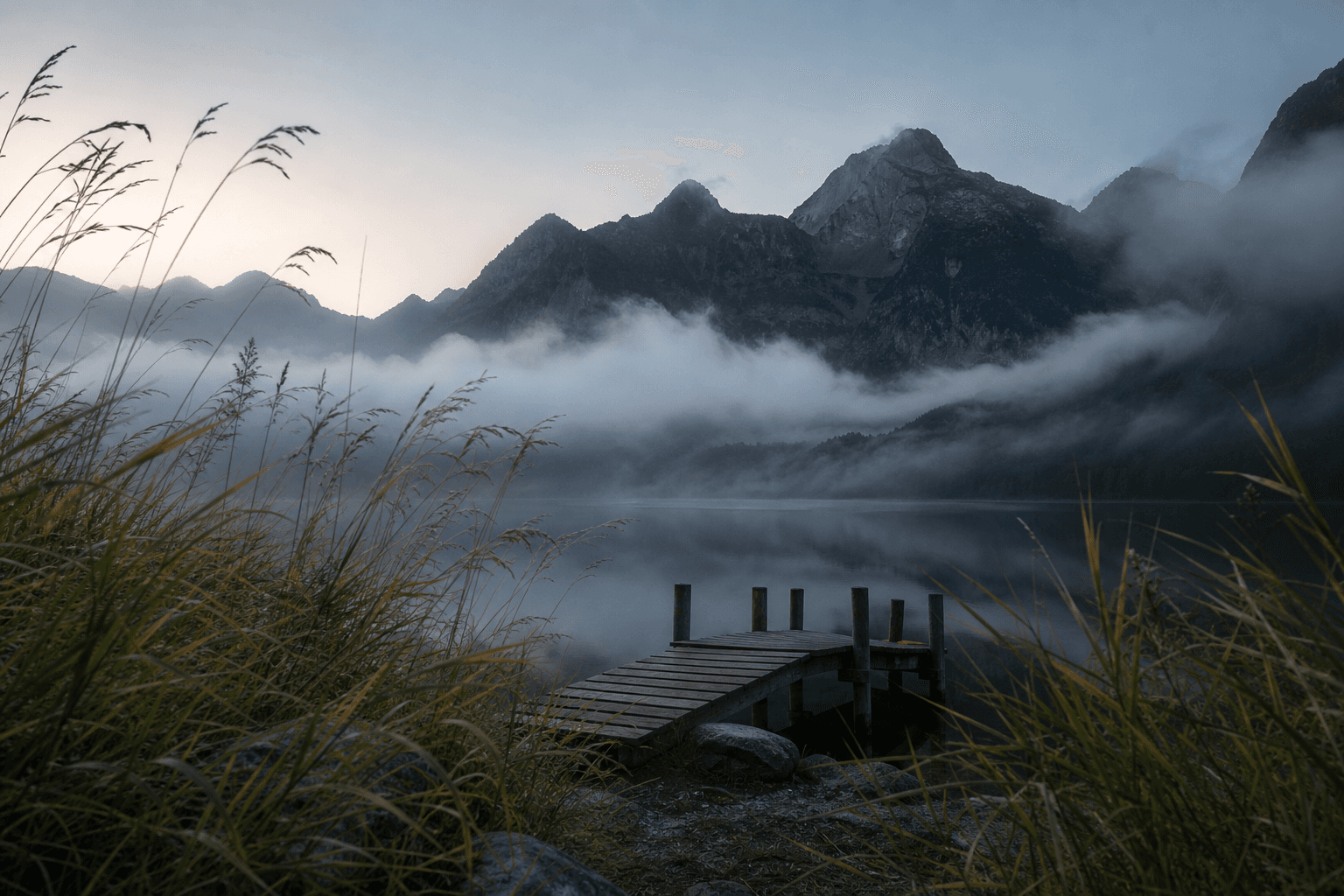 Misty alpine lake, wooden dock, wild grass, and dark mountain peaks at dawn.